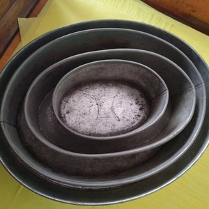 K&ouml;nnte beinhalten: Ein Satz von vier ovalen Backformen aus Metall, die ineinander verschachtelt sind. Die gr&ouml;&szlig;te Form bildet die Basis, mit zunehmend kleineren Formen im Inneren. Das Metall hat ein verwittertes, gealtertes Aussehen.