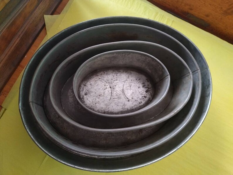 K&ouml;nnte beinhalten: Ein Satz von vier ovalen Backformen aus Metall, die ineinander verschachtelt sind. Die gr&ouml;&szlig;te Form bildet die Basis, mit zunehmend kleineren Formen im Inneren. Das Metall hat ein verwittertes, gealtertes Aussehen.