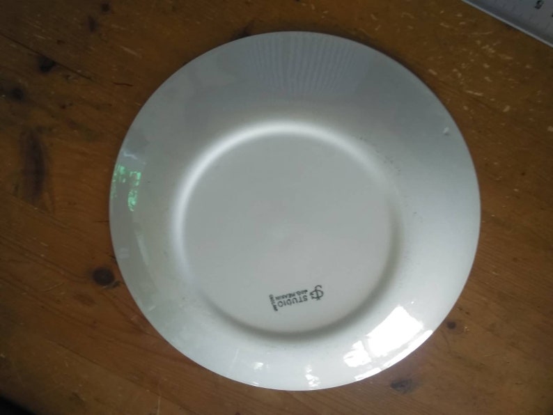 K&ouml;nnte beinhalten: Ein wei&szlig;er Keramikteller mit einem erh&ouml;hten Rand und einer dezenten strukturierten Oberfl&auml;che. Der Teller hat ein kleines Logo in der Mitte, das "Studio Nova" lautet.