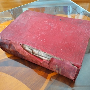 May include: A red hardcover book with a worn and damaged spine. The book has a decorative pattern on the cover and is slightly faded.