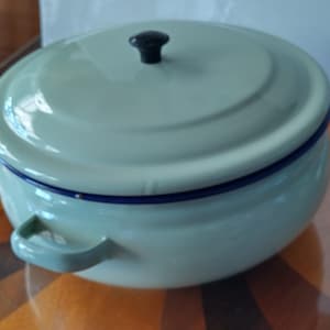 May include: A round, light mint green enamel pot with a matching lid. The pot features two side handles and a dark blue rim. A small, black, circular knob is centred on the lid. The finish is smooth and glossy, with a simple, functional design.