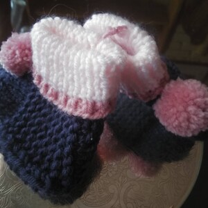 May include: A pair of knitted baby booties with pink and blue yarn. The booties have a pink pom-pom on each side.