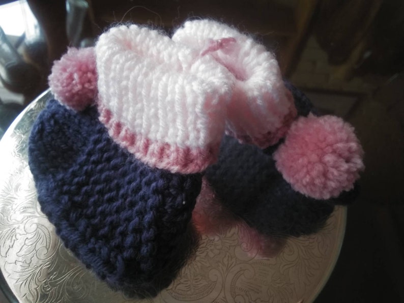 May include: A pair of knitted baby booties with pink and blue yarn. The booties have a pink pom-pom on each side.