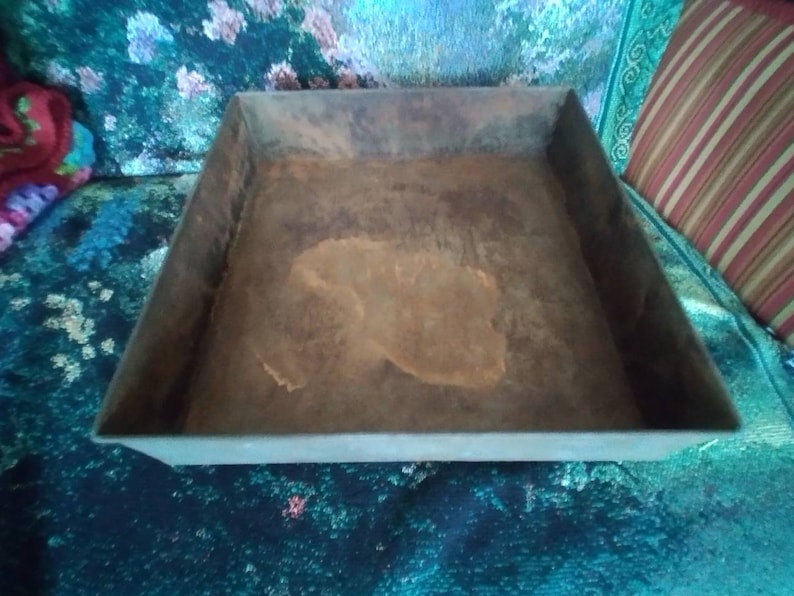 May include: A square metal tray with a rusty brown surface. The tray is empty and has a slightly raised edge.