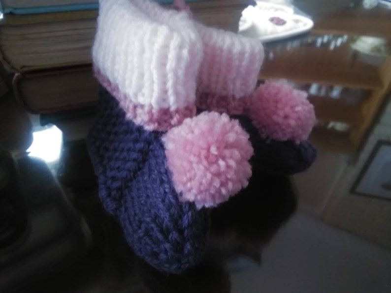 May include: A pair of knitted baby booties with pink pom-poms. The booties are purple and white with a pink stripe.