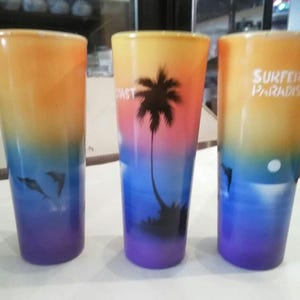 May include: Three colorful glass shot glasses with a beach scene design. The glasses are decorated with a sunset, palm trees, and dolphins. One glass has the text "Surfers Paradise" printed on it.
