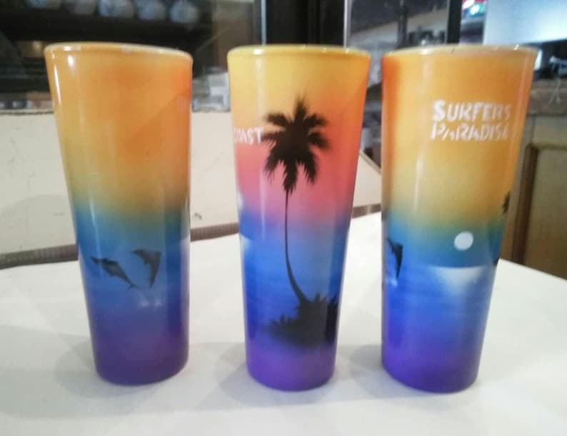 May include: Three colorful glass shot glasses with a beach scene design. The glasses are decorated with a sunset, palm trees, and dolphins. One glass has the text "Surfers Paradise" printed on it.