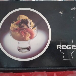 Puede incluir: Una caja negra y blanca con la palabra "REGIS" impresa en blanco. La caja presenta una imagen de primer plano de un cuenco de vidrio transparente lleno de una ensalada de mariscos. La ensalada incluye camarones, cangrejo y otros mariscos.