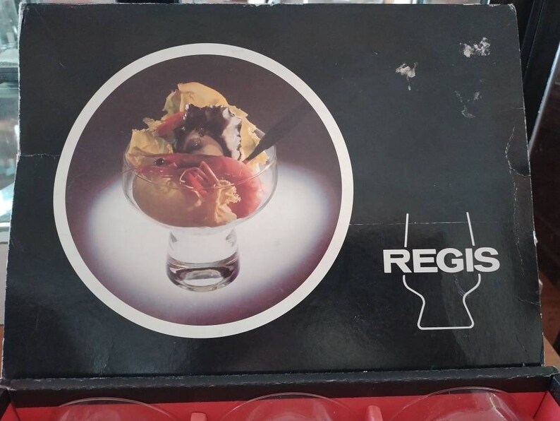 Puede incluir: Una caja negra y blanca con la palabra "REGIS" impresa en blanco. La caja presenta una imagen de primer plano de un cuenco de vidrio transparente lleno de una ensalada de mariscos. La ensalada incluye camarones, cangrejo y otros mariscos.