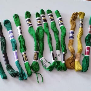 May include: A collection of eleven skeins of embroidery floss in various shades of green and gold. Each skein is secured with a paper label featuring the brand name Semco, colour numbers, and the length of 7.3 metres printed on the paper bands.