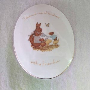 May include: White ceramic plate with a gold rim featuring a whimsical illustration of a bunny, two mice, and a butterfly. The plate has the text "Share a cup of kindness with a friend..." in gold lettering.