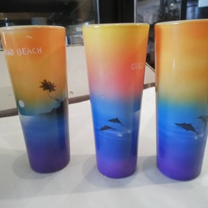 May include: Three colorful glass shot glasses with a sunset and dolphin design. The glasses are decorated with a gradient of orange, yellow, blue, and purple. The text "Road Beach" is printed on one glass, and the text "Gold" is printed on another glass.