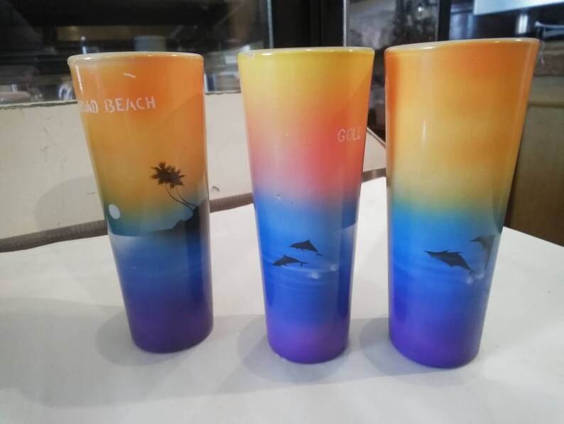 May include: Three colorful glass shot glasses with a sunset and dolphin design. The glasses are decorated with a gradient of orange, yellow, blue, and purple. The text "Road Beach" is printed on one glass, and the text "Gold" is printed on another glass.