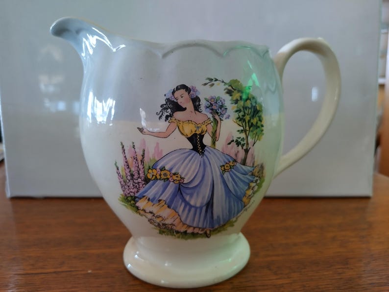 以下が含まれることがあります： 青いドレスを着た女性が花を持っている、青と黄色の花柄の白い陶器のピッチャー。ピッチャーは波打つ縁と取っ手があります。