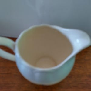 以下が含まれることがあります： 持ち手付きの白磁のピッチャーで、薄い青色の釉薬がかかっています。ピッチャーは空で、滑らかで丸みを帯びた形をしています。