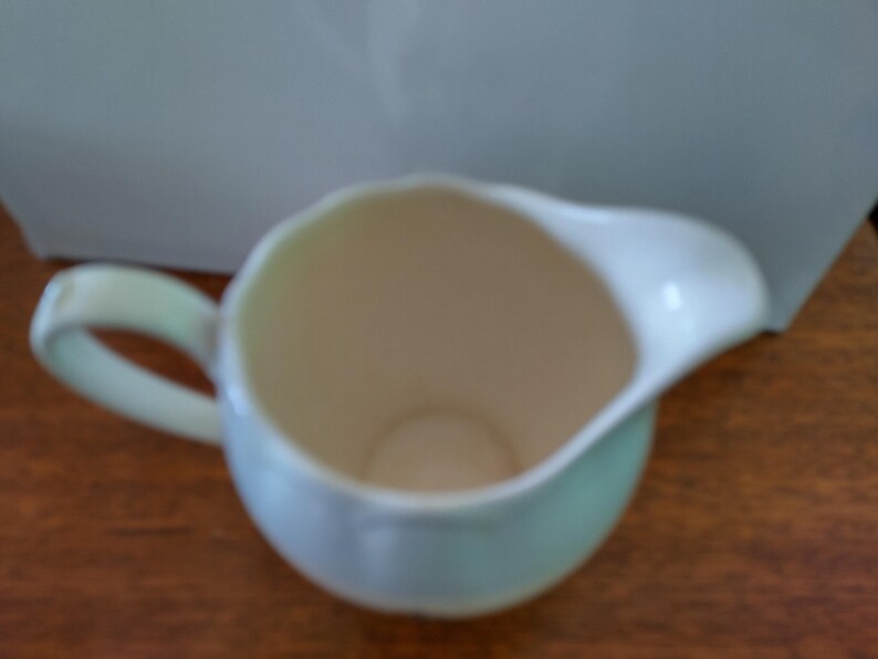 以下が含まれることがあります： 持ち手付きの白磁のピッチャーで、薄い青色の釉薬がかかっています。ピッチャーは空で、滑らかで丸みを帯びた形をしています。