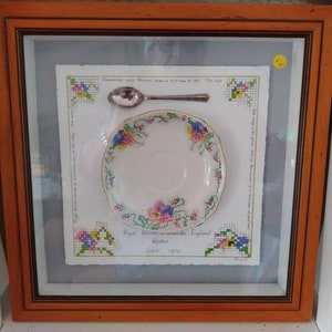 May include: A framed piece of cross-stitch embroidery featuring a small teacup and saucer with a floral design. The embroidery is surrounded by a white border and a light blue background. The text "Royal Winton commemorative England" and "Carol 1994" is included in the embroidery.