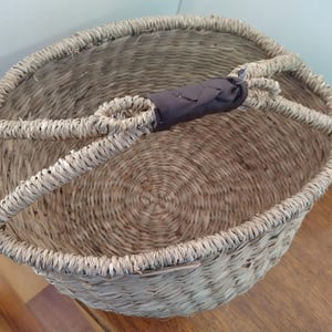 May include: A woven basket with a rounded shape and a dark gray handle. The basket is made of natural fibers, with a light brown color and a textured surface. The handle is made of a woven fabric. The basket is empty.