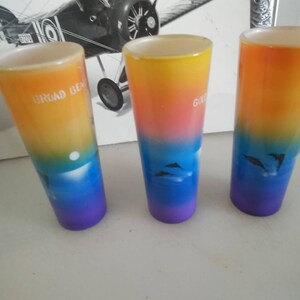 May include: Three colorful glass shot glasses with a gradient design. The glasses are decorated with a sunset scene and dolphins. The text on the glasses reads "Broad Beach", "Grand Cayman", and "South Padre Island".