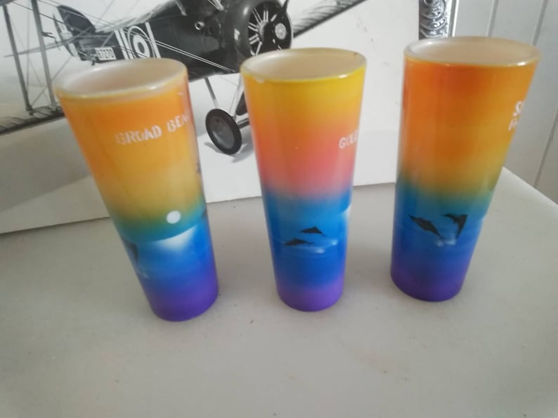 May include: Three colorful glass shot glasses with a gradient design. The glasses are decorated with a sunset scene and dolphins. The text on the glasses reads "Broad Beach", "Grand Cayman", and "South Padre Island".
