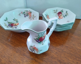 May include: A set of white ceramic dishes with a green rim and a floral pattern featuring fruits and berries. The set includes a large octagonal bowl, a smaller octagonal bowl, and a pitcher.