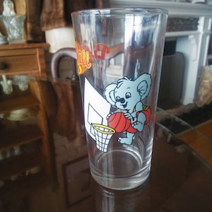 May include: A clear glass with a cartoon koala playing basketball. The koala is wearing a red and yellow jersey and holding a basketball. The glass has the word "Ball" printed on it.
