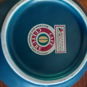 Puede incluir: La parte inferior de una tetera de cer&aacute;mica azul turquesa con un borde blanco. La tetera tiene una pegatina que dice "Cafe Culture" con una imagen de grano de caf&eacute; y "Microwave and Dishwasher Safe Porcelain". La pegatina tambi&eacute;n tiene el logotipo "Maxwell Williams Designer Homewares".