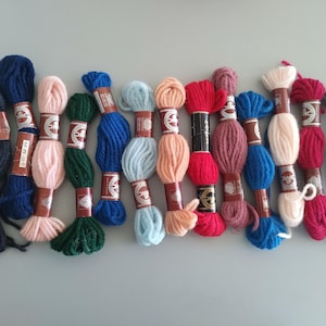 May include: A selection of colourful embroidery floss skeins in shades of blue, pink, green, orange, red, and purple. Each skein is wrapped with a paper label. The floss is arranged on a light grey surface.