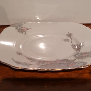 May include: A white, scalloped-edge plate with a floral design in pink, grey, and blue. The plate has a gold trim and a slightly oval centre. The floral pattern is arranged around the edge of the plate.