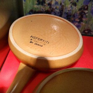 May include: A brown ceramic casserole dish with a white rim. The bottom of the dish has the text "NEFERTITI STONEWARE BY DIANA" printed on it.