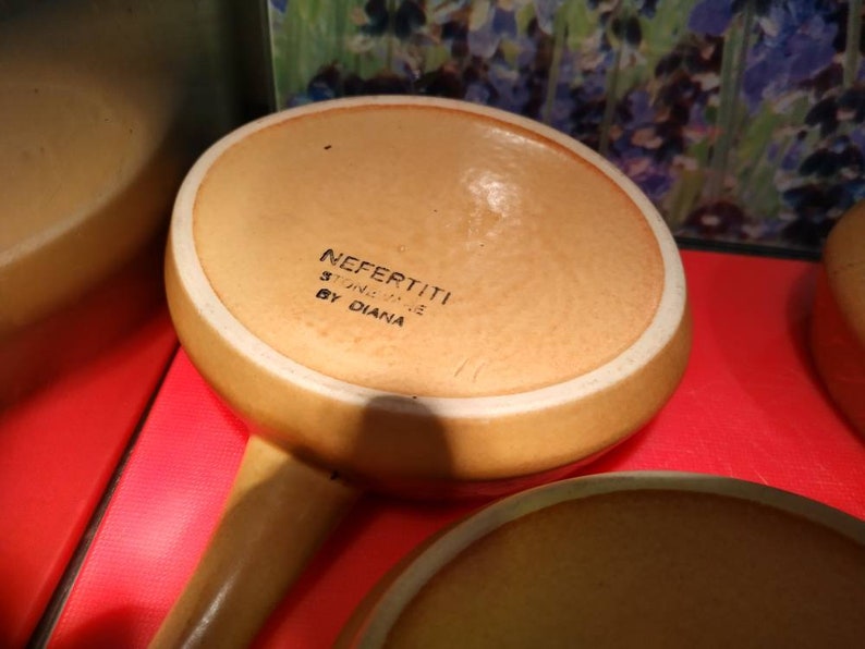 May include: A brown ceramic casserole dish with a white rim. The bottom of the dish has the text "NEFERTITI STONEWARE BY DIANA" printed on it.