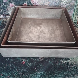 May include: Three square metal baking pans, nested together. The pans are silver in color and have a worn, rustic look.