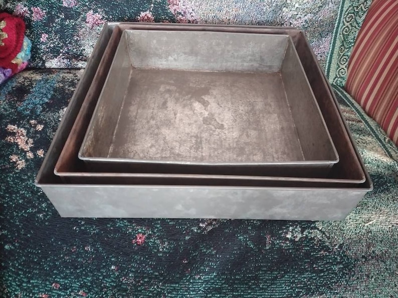 May include: Three square metal baking pans, nested together. The pans are silver in color and have a worn, rustic look.