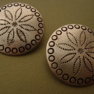 May include: Two silver-toned metal buttons with a decorative design. Each button features a circular border with a raised, dotted pattern. The centre of each button has a stylised flower design with a dotted outline.