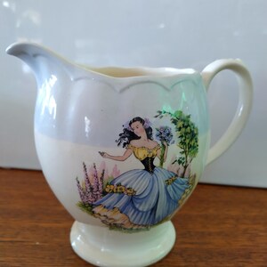 以下が含まれることがあります： 青いドレスと黄色のコルセットを着た女性が描かれた花柄の白い陶器のクリーマー。クリーマーは波打つ縁と足付きのベースを持っています。