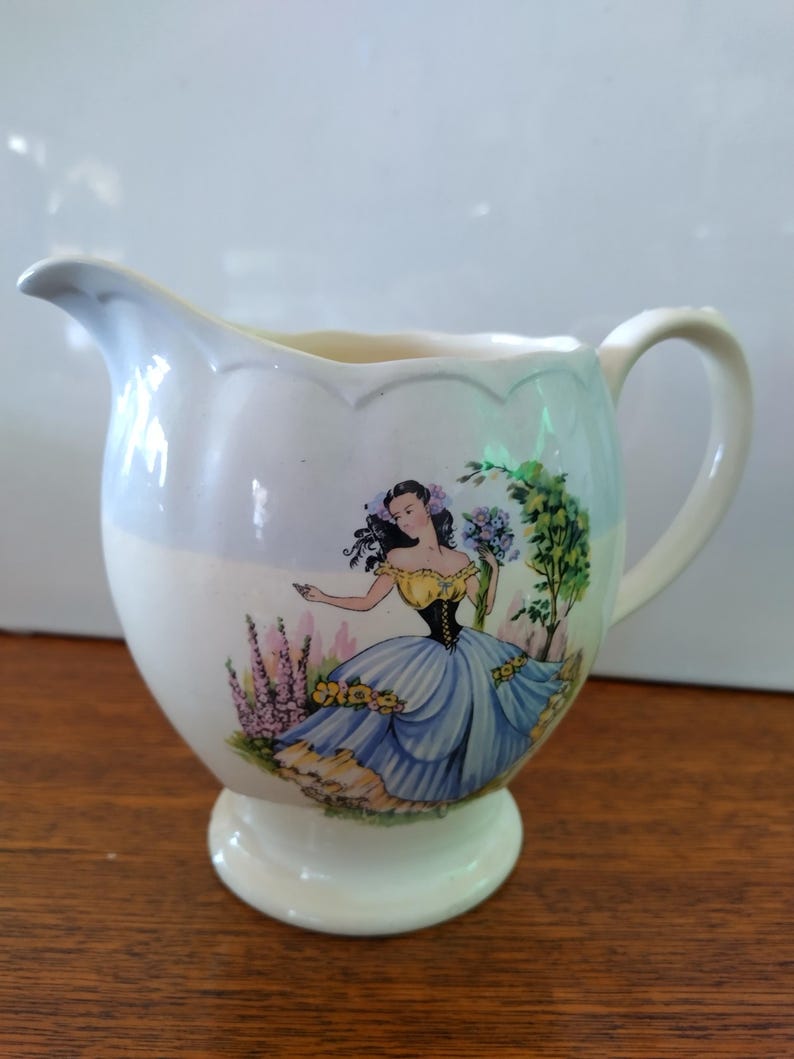 以下が含まれることがあります： 青いドレスと黄色のコルセットを着た女性が描かれた花柄の白い陶器のクリーマー。クリーマーは波打つ縁と足付きのベースを持っています。