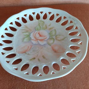 May include: A white porcelain decorative plate with a delicate openwork design. The plate features a hand-painted floral design with a pink rose and green leaves.