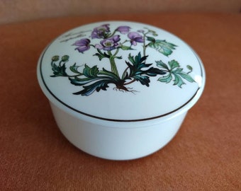 May include: A white porcelain trinket box with a floral design. The lid features a delicate illustration of a white flower with green leaves. The box has a gold trim around the edge.