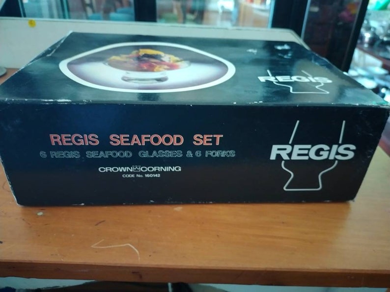 Puede incluir: Una caja negra con el texto "REGIS SEAFOOD SET 6 REGIS SEAFOOD GLASSES & 6 FORKS CROWN CORNING CODE No 160142 REGIS" impreso en ella. La caja contiene un juego de seis vasos para mariscos.