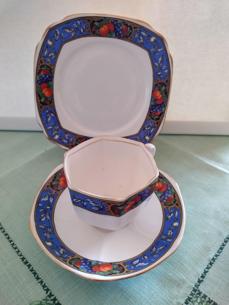 Puede incluir: Un juego de taza y platillo de t&eacute; blanco con un borde azul y dorado y un dise&ntilde;o de frutas. El platillo es de forma cuadrada y la taza es octogonal.