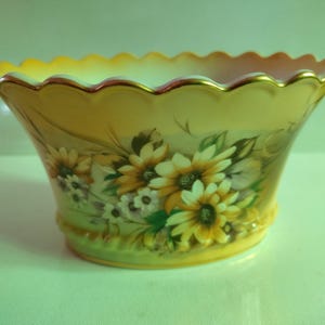 May include: A decorative ceramic bowl with a scalloped rim and a floral design. The bowl is painted with yellow, white, and green flowers on a cream background.
