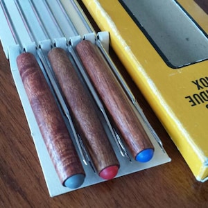 May include: Six fondue forks with wooden handles in a white cardboard box. The box has a yellow label that reads "6 GARFOS PARA FONDUE EM INOX".