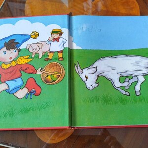 以下が含まれることがあります： 左ページにはリンゴを持った少年と豚が描かれたイラスト入りの本。右ページには緑の草原を走る白いヤギが描かれており、上部に「caprella」という文字が書かれている。