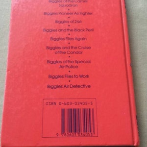 May include: A red book with a list of titles from the "Biggles" series. The titles include "The Boy Biggles", "Biggles of the Camel Squadron", and "Biggles Pioneer Air Fighter". The book has an ISBN and item code.