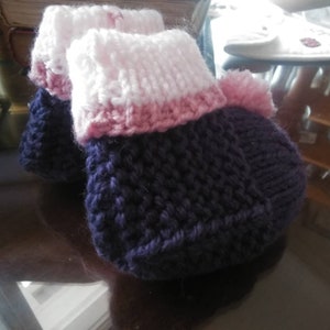 May include: A pair of knitted baby booties in purple, pink, and white. The booties have a textured knit pattern and a fluffy pom-pom on top.