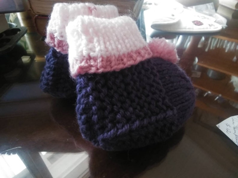May include: A pair of knitted baby booties in purple, pink, and white. The booties have a textured knit pattern and a fluffy pom-pom on top.