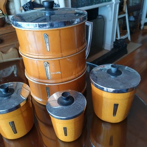 May include: Set of four vintage kitchen canisters with chrome lids. The canisters are brown with a woodgrain pattern and have labels that say "Sugar", "Flour", "Tea", and "Coffee".