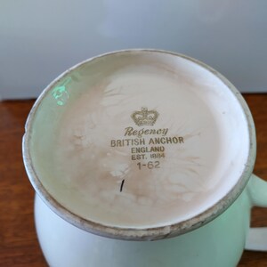 以下が含まれることがあります： 緑色の取っ手が付いた白い陶器製のマグカップ。マグカップの底には、"Regency British Anchor England Est. 1884 1-62"と刻まれた金色のロゴがあります。