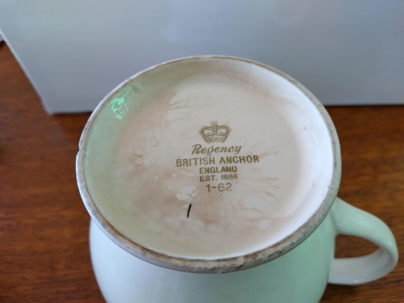 以下が含まれることがあります： 緑色の取っ手が付いた白い陶器製のマグカップ。マグカップの底には、"Regency British Anchor England Est. 1884 1-62"と刻まれた金色のロゴがあります。