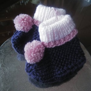 May include: A pair of knitted baby booties with pink and purple yarn. The booties have a pink pom-pom on the top.